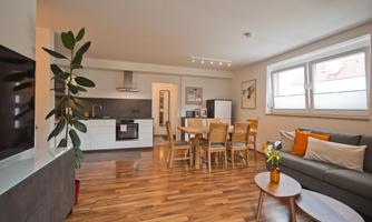 A modern living room with an open kitchen and a dining area. The decor is bright and friendly, with wooden flooring and stylish furniture.