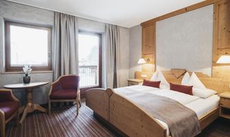 A cozy bedroom with a wooden bed and soft pillows. There is a table with chairs and a window that lets in daylight.