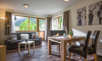An inviting living room with large windows and a view of the mountains. There is a cozy couch and a wooden table with chairs.