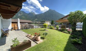 A green garden landscape with flowers and a view of high mountains. The sky is clear and the sun is shining.