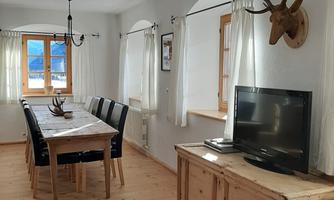 A bright dining room with a long wooden table and black chairs. A deer antler hangs on the wall and a television stands on a wooden sideboard.