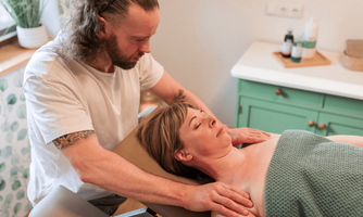 A man gives a woman a massage in a warm, friendly room. A blanket is on the table, and in the background, care products are visible.