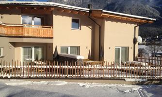 A modern house in the snow, surrounded by mountains. The garden is fenced and the sun is shining on the terrace.