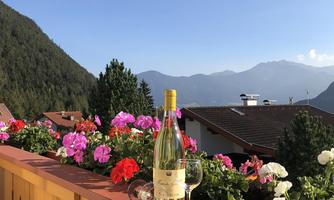 A terrace with colorful flowers and a view of the mountains. A bottle of wine and a glass are ready for enjoyment.