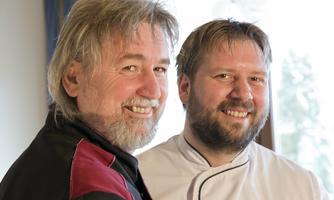 Two smiling men are standing next to each other. One is wearing a chef's jacket, while the other has a black top.