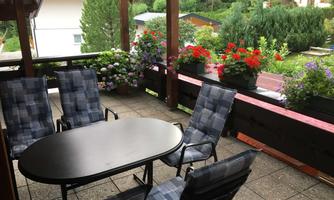 A cozy terrace with a round table and several chairs.  
Surrounded by colorful flowers and a green landscape.