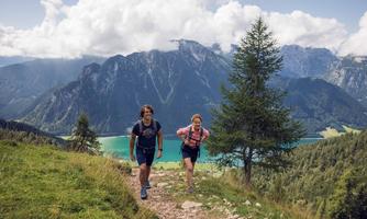 Two hikers on a narrow path in the mountains. In the background, impressive mountains and a sparkling lake can be seen.