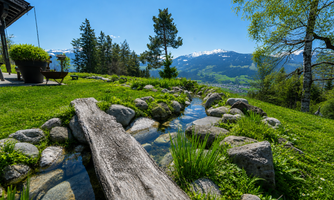 A picturesque landscape with a small stream surrounded by stones. In the background, green hills and snow-capped mountains can be seen.