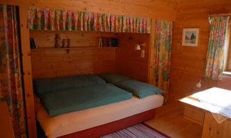 A cozy wooden bedroom with a double bed and colorful curtains. A lamp is on the nightstand, and there is a small desk window.