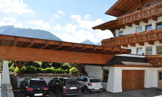 A modern house with a wooden roof and several cars underneath. In the background, mountains and a blue sky can be seen.