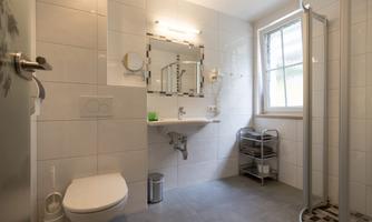 A modern bathroom with a shower, a toilet, and a sink. The walls are tiled in light colors, and there is natural lighting from a window.