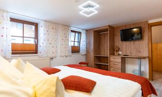 A cozy bedroom with a double bed and colorful cushions. The room features wooden paneling, a television, and windows with curtains.