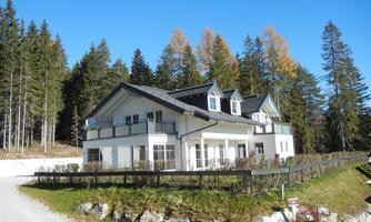 A modern house in a rural setting, surrounded by trees and a green meadow. The sky is clear and blue.