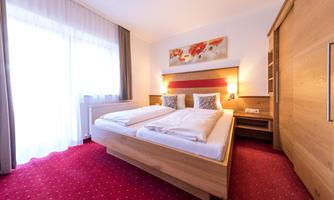A cozy bedroom with a bright ambiance. There is a large bed, wooden shelves, and plenty of daylight through the window.