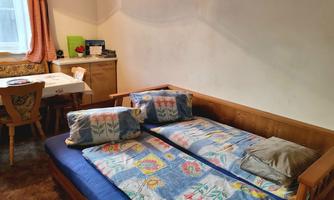 A simple room with a wooden bed and colorful cushions. In the background, there is a table with chairs and a desk space.