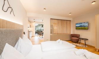 A modern hotel room with two beds, a television, and an armchair. The room has large windows and a warm wooden floor.