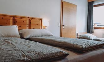 A simple bedroom with two single beds and a wood-paneled headboard. The window lets in natural light and the walls are painted in a light color.