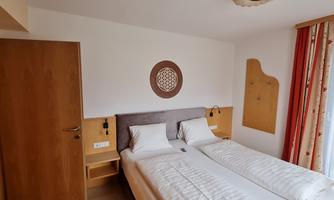 A cozy room with two single beds and a simple wooden wardrobe.  
On the wall, there is a decorative wall design and large windows with curtains.