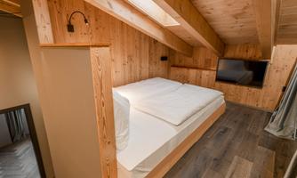 A modern bedroom with wood-paneled walls and a large window. The bed is simple and inviting, complemented by a television on the wall.