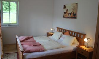 A cozy bedroom with a double bed and wooden dressers. The room has a window with a view of greenery and a picture on the wall.