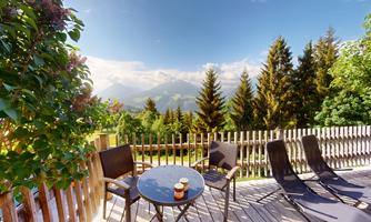 A cozy balcony with a view of the mountains and trees. Two comfortable seating areas and a table are ready.