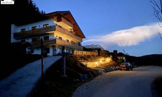 A cozy, illuminated building in a rural setting at dusk. The path gently leads to the front door and some trees are visible in the background.