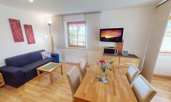 A cozy living space with a blue sofa and a wooden dining table. Decorative pictures hang on the wall, and a television is visible.