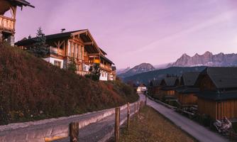 A picturesque mountain village with wooden chalets and a gentle path. In the background, majestic mountains and a beautiful, colorful sky can be seen.