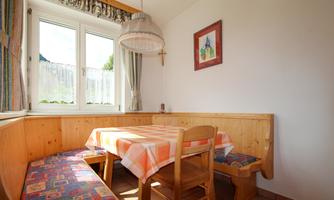 A cozy dining area with a wooden table and benches. Bright windows let in plenty of light and a decorative lamp hangs over the table.