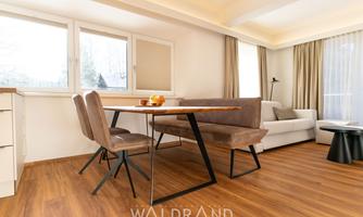 A modern living room with a dining area and a comfortable sofa. Large windows provide plenty of daylight and a friendly atmosphere.