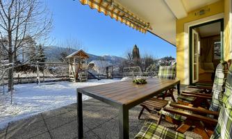 A cozy terrace with a wooden table and comfortable chairs. In the background, there is a snow-covered garden and the mountains.