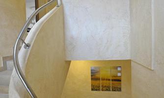 A bright staircase with a curved railing and a pot with a plant on top. A picture of a landscape hangs on the wall.