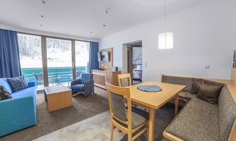 A modern living room with a blue sofa, a table, and chairs. Large windows offer a view of the snowy landscape outside.
