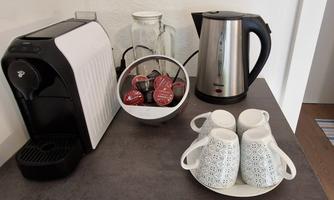 A cozy kitchen corner with a coffee machine, a kettle, and a bowl of coffee capsules. Two decorative cups are placed on a saucer.