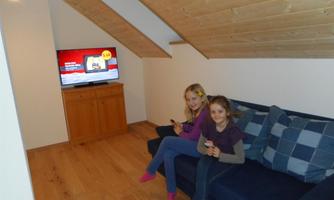 A cozy room with wooden ceilings and a sofa. Two girls are sitting on the sofa and watching TV.