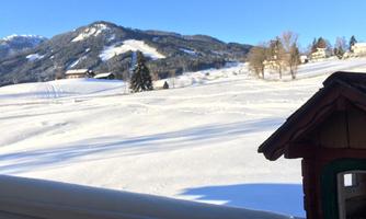A snowy landscape with gentle hills and mountains in the background. In the foreground stands a small wooden house.