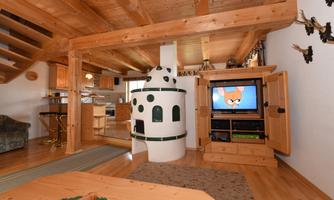 A cozy living space with wooden beams and a fireplace. In the background, a modern kitchen and a television are visible.