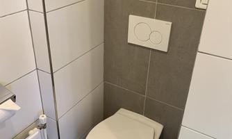 A modern bathroom with a simple toilet in neutral tones. Next to the toilet is a small heater and a toilet paper roll.
