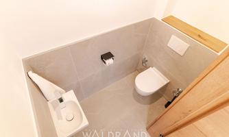 A modern bathroom with a toilet, a hand basin, and gray tiles. The design is minimalist and bright.