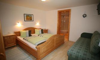 A cozy bedroom with a wooden double bed and a green sleeping couch. The walls are painted light, and there is a small table and a picture on the wall.