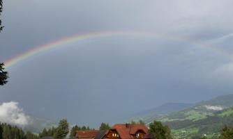A farmhouse in a green landscape under a colorful rainbow. The sky is overcast, and the mountains are visible in the background.