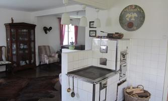 A cozy kitchen with a traditional stove and modern furniture. In the background, there is a living area with a large cabinet and a comfortable armchair.