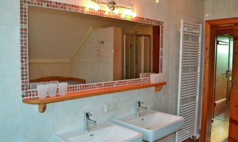 A modern bathroom with two sinks and a large mirror. The walls are tiled in light colors, and there is a shower in the background.