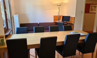A bright dining area with a long wooden table and black chairs. In the background, there is a sofa and a monitor.