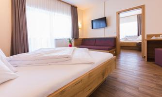A modern hotel room with a double bed, television, and a cozy seating area. Large windows provide plenty of daylight.
