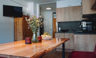 A modern kitchen with a wooden desk and a bouquet of flowers in a vase. In the background, kitchen cabinets and a television are visible.