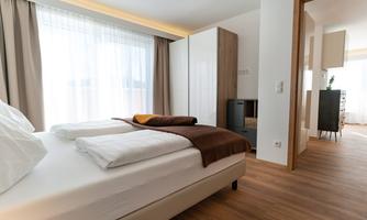 A modern bedroom with a large bed and light wooden floor. Large windows provide plenty of light and a welcoming atmosphere.