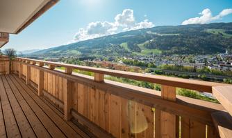 A beautiful balcony with a wooden railing and a view of green mountains. The sun is shining, and the sky is clear.
