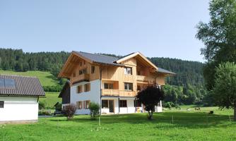 A modern wooden house with two floors stands in a green landscape. In the background, gentle hills and trees can be seen.