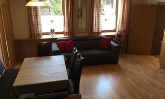 A bright living room with a sofa, red cushions, and a table. The large windows let in plenty of daylight.
